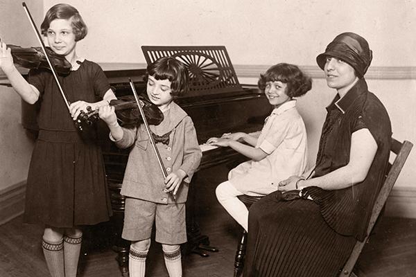 戴着帽子的女人带着孩子在弹钢琴，两个孩子在拉小提琴